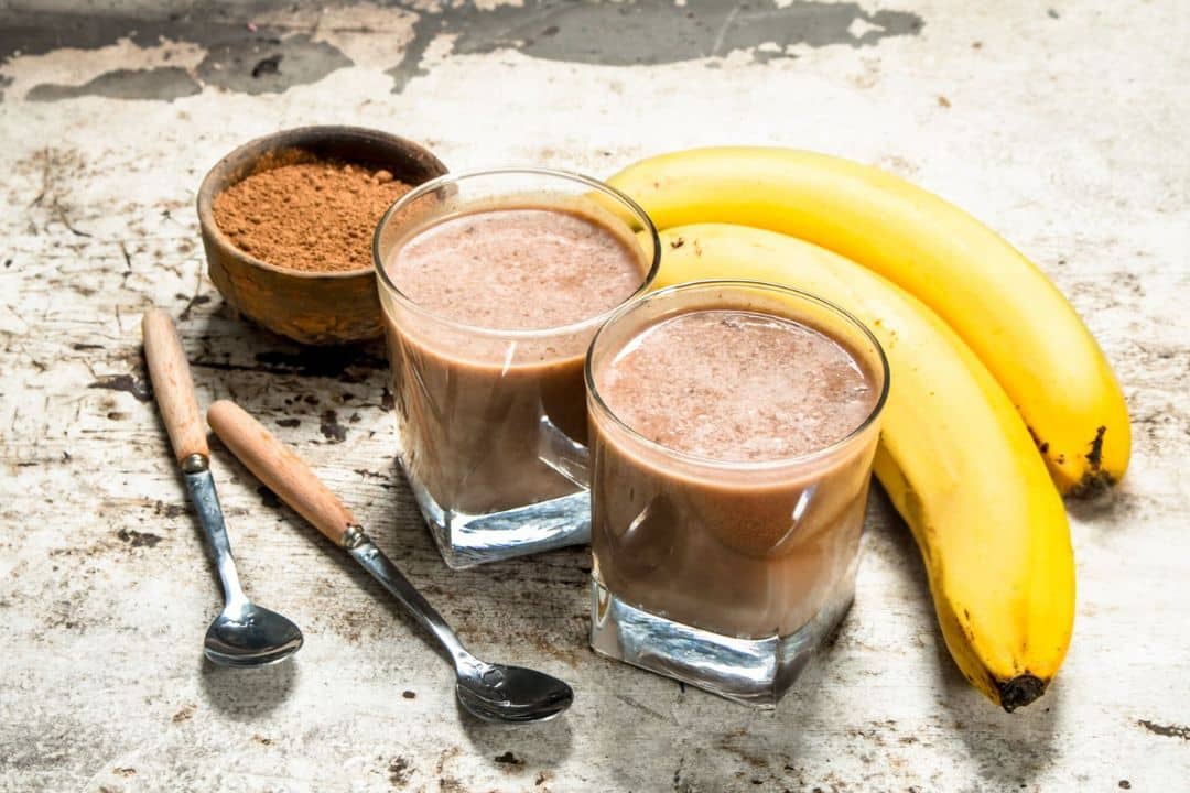 A rustic setup featuring two glasses of a creamy chocolate-banana protein shake, made with Primabolics Iso-Ripped Caramel White Chocolate, surrounded by fresh bananas, a small bowl of protein powder, and two spoons with wooden handles. The image conveys a nutritious and delicious start to the day.