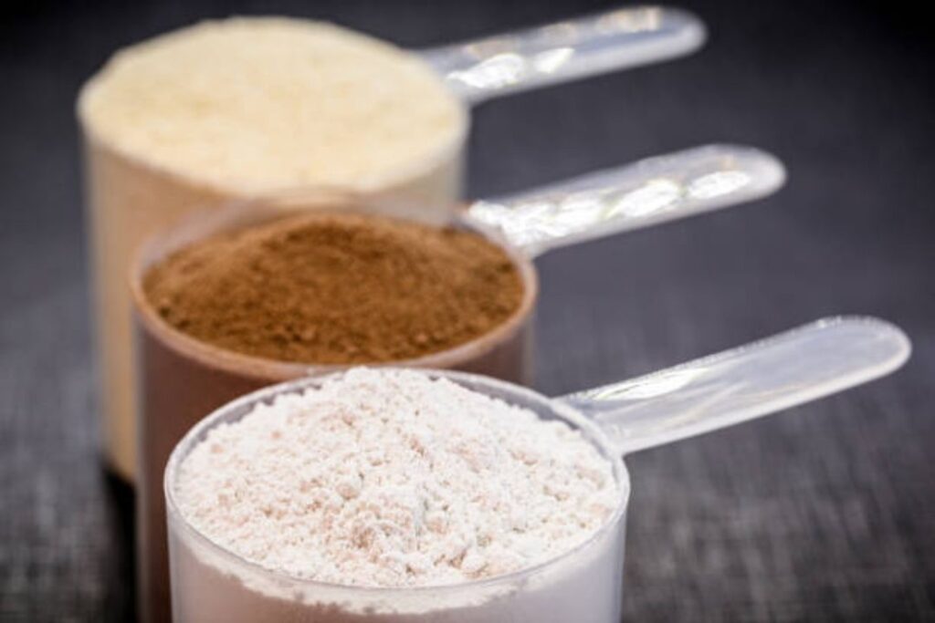 Three scoops of protein powder in different colors (white, brown, and off-white) arranged in a row with clear plastic scoops on a dark surface.